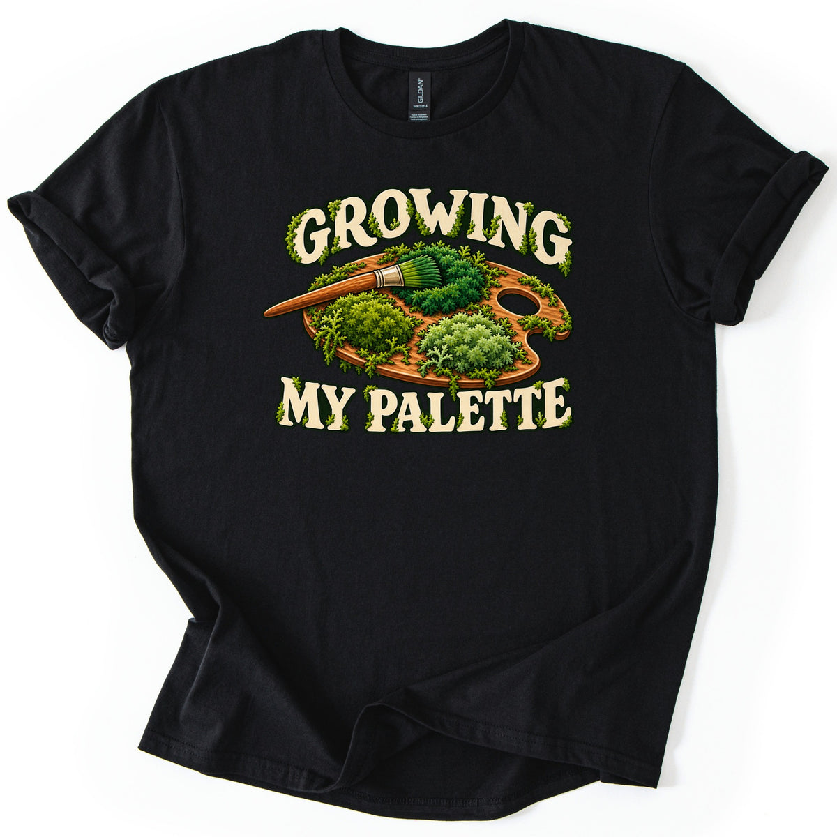 Growing My Palette Comfort Colors T-shirt in Black, laid flat. An artist's wooden palette and paintbrush covered in green moss and plants, between the words Growing My Palette in cream letters edged with moss. The sleeves are rolled on a white background.