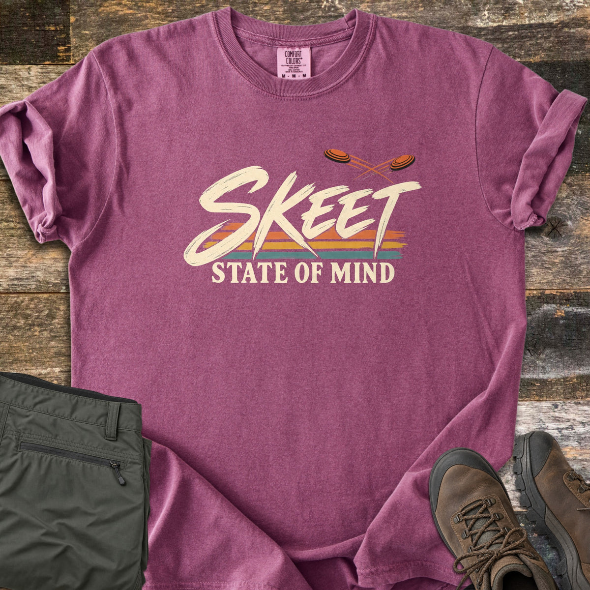 Skeet State of Mind Comfort Colors T-shirt in Berry, laid flat. Brushy cream letters spell Skeet over orange, yellow and teal stripes, with two orange clay targets flying above and State of Mind below. Olive pants and brown hiking boots sit on rustic wood planks.
