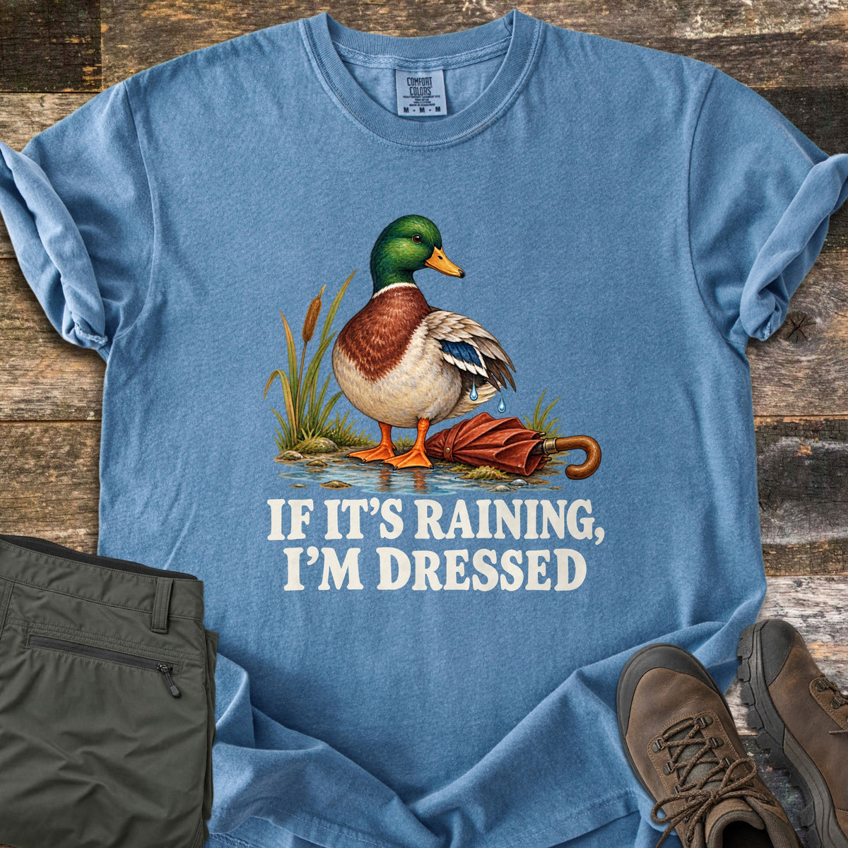If It's Raining, I'm Dressed Comfort Colors T-shirt in Blue Jean, laid flat. A mallard duck stands in a puddle by cattails, water dripping off its wing, next to a closed red umbrella, above the words If It's Raining, I'm Dressed in white. Olive pants and brown hiking boots lie on weathered wood.