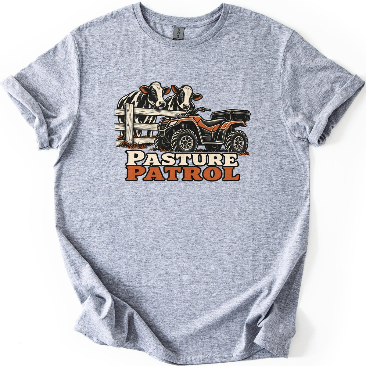 Pasture Patrol Comfort Colors T-shirt in Sport Grey, laid flat. Two black and white cows look over a wooden fence beside an orange ATV with a rear cargo box, above the words Pasture Patrol. The shirt lies on a white background with its sleeves rolled.
