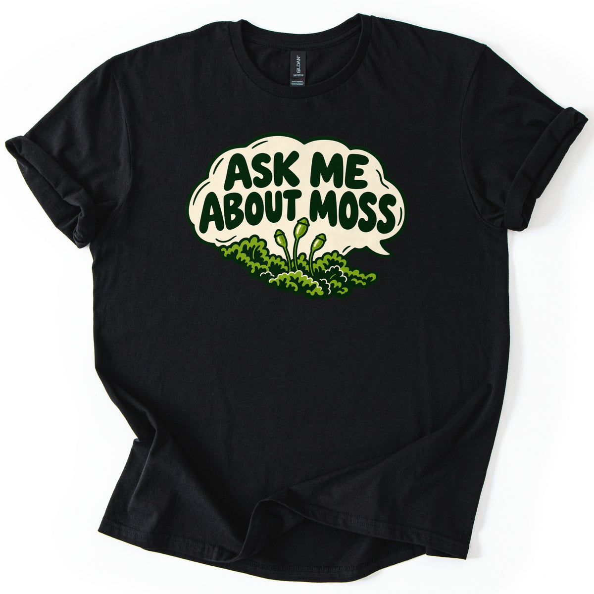 Ask Me About Moss Comfort Colors T-shirt in Black, laid flat. A cream speech bubble with ASK ME ABOUT MOSS in bold dark green letters sits over a cartoon mound of green moss with three thin spore stalks. The sleeves are rolled and the shirt lies on a white surface.