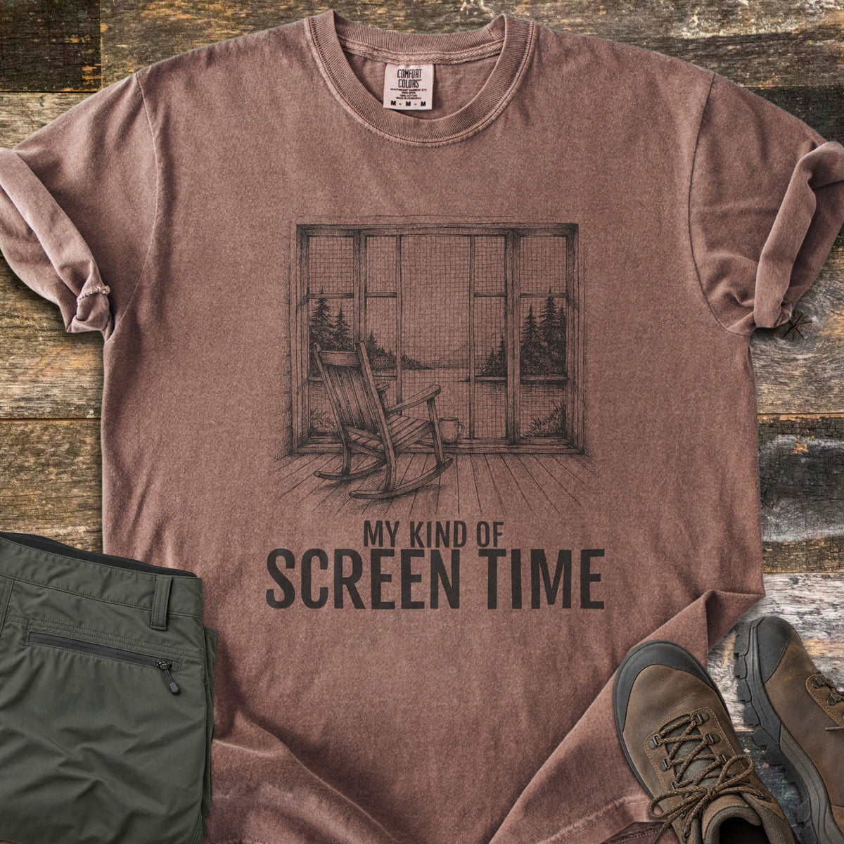 My Kind Of Screen Time Comfort Colors T-shirt in Espresso, laid flat. A black sketch of a wooden rocking chair and mug facing a screened porch window with a view of a lake and pine trees, above the words My Kind Of Screen Time. Folded olive hiking pants and brown hiking boots sit on rustic wood.