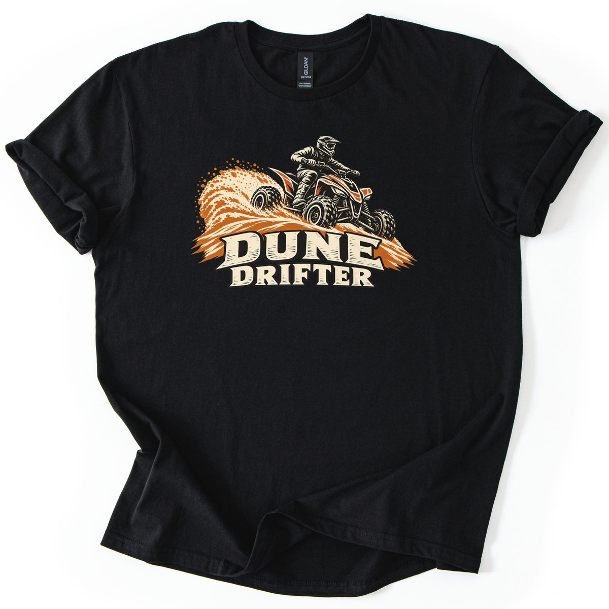 Dune Drifter Comfort Colors T-shirt in Black, laid flat. A helmeted rider on an orange and black ATV sprays orange sand over a dune above the words Dune Drifter in cream letters. The shirt has cuffed sleeves and lies on a white background.