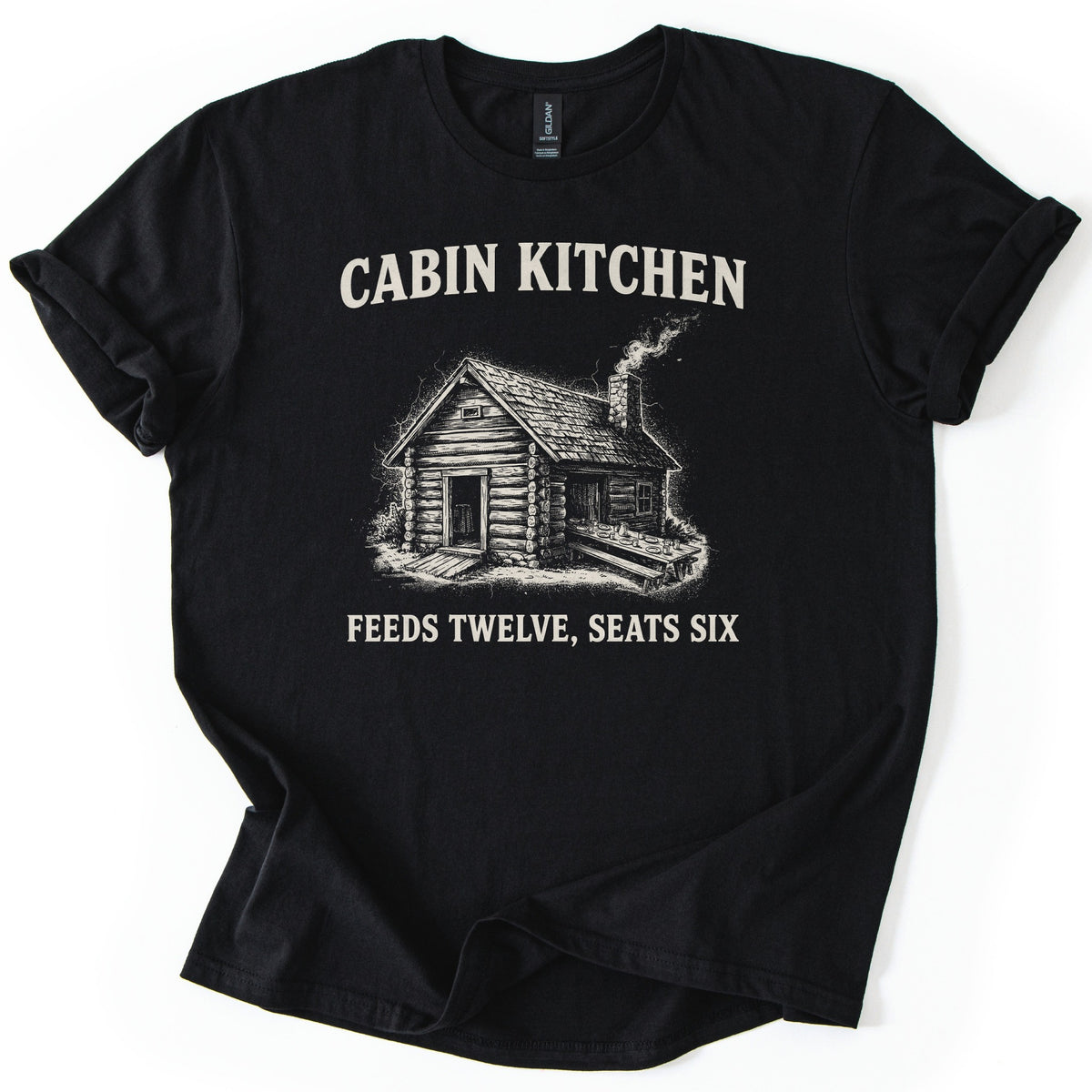 Cabin Kitchen Comfort Colors T-shirt in Black, laid flat. Cream text reads Cabin Kitchen above a sketched log cabin with a smoking chimney and a long outdoor table set with dishes, with Feeds Twelve, Seats Six below. Sleeves are cuffed on a white background.