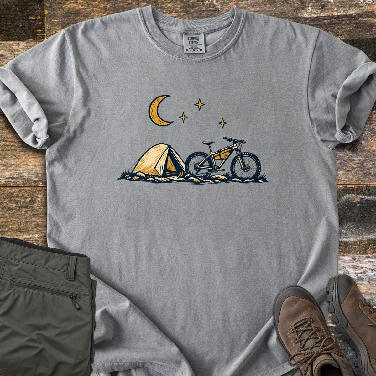 Bikepacking Under the Stars Comfort Colors T-shirt in Grey, laid flat. A yellow tent and a mountain bike with a frame bag sit on grassy ground under a crescent moon and three stars, with no words. Olive hiking pants and brown boots lie beside it on weathered wood planks.