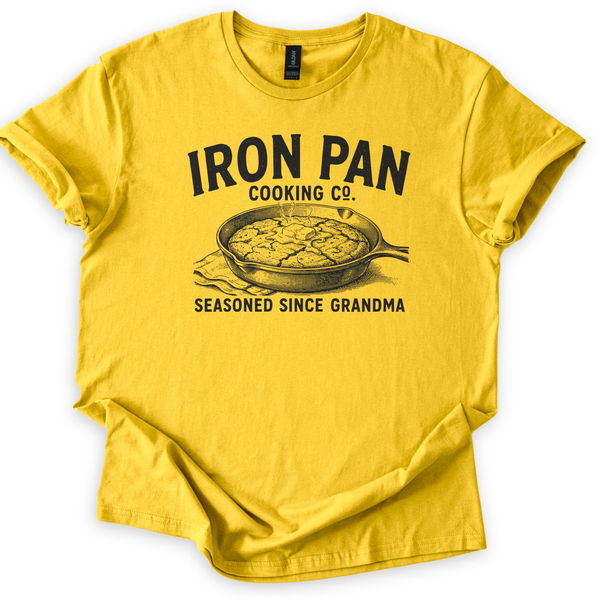 Iron Pan Cooking Co Comfort Colors T-shirt in Daisy, laid flat. Black print of the words Iron Pan Cooking Co. above an engraved-style cast iron skillet of cornbread topped with melting butter on a cloth, with Seasoned Since Grandma below. Cuffed sleeves on a white background.