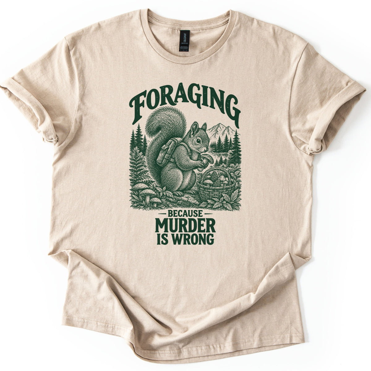 Foraging Murder Squirrel Comfort Colors T-shirt in Sand, laid flat. Foraging arches over a dark green etched squirrel wearing a backpack and holding a mushroom beside a basket of mushrooms, with pines and a mountain behind. Below: Because Murder Is Wrong. Cuffed sleeves, white background.