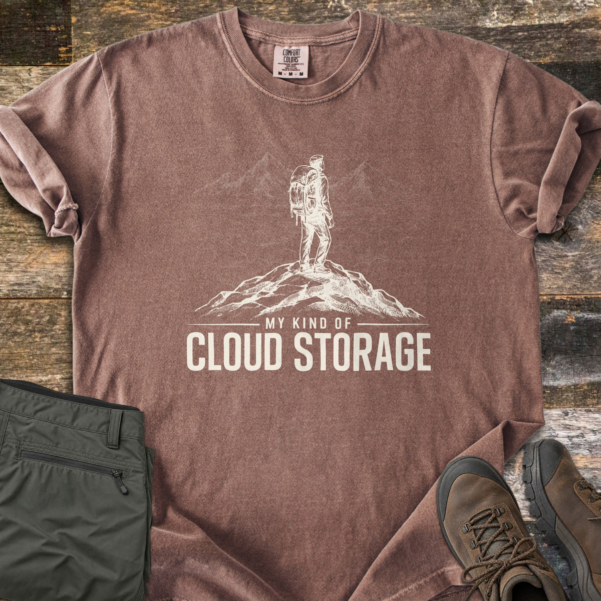 My Kind Of Cloud Storage Comfort Colors T-shirt in Espresso, laid flat. A cream sketch of a backpacker on a rocky summit facing peaks above the clouds, over the words My Kind Of Cloud Storage. Sleeves are rolled, with olive hiking pants and brown hiking boots beside it on weathered wood.