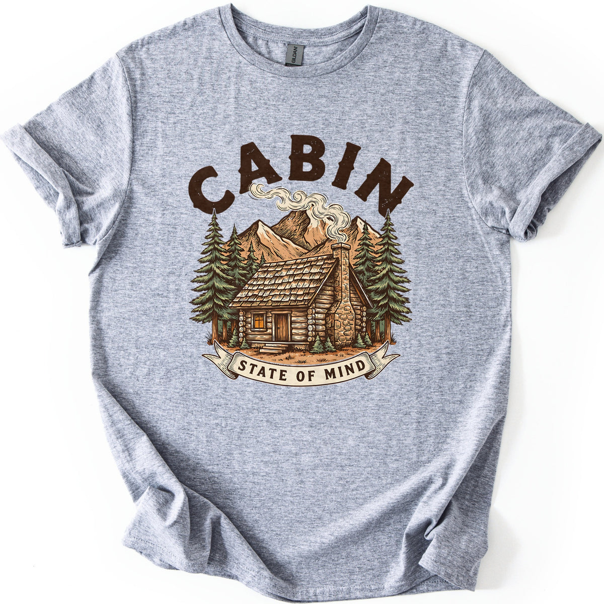 Cabin State Of Mind Comfort Colors T-shirt in Sport Grey, laid flat. CABIN arches in distressed dark brown letters over a log cabin with a smoking stone chimney, pine trees and mountains, above a banner reading STATE OF MIND. The shirt lies on white with cuffed sleeves.