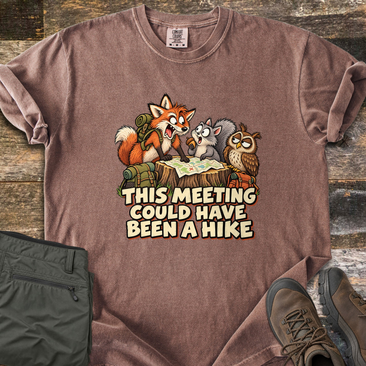 This Meeting Could Have Been a Hike Comfort Colors T-shirt in Espresso, laid flat. A cartoon fox with a backpack, a wide-eyed squirrel and a bored owl look over a trail map on a stump above the words This Meeting Could Have Been a Hike. Folded olive pants and hiking boots sit on dark wood.
