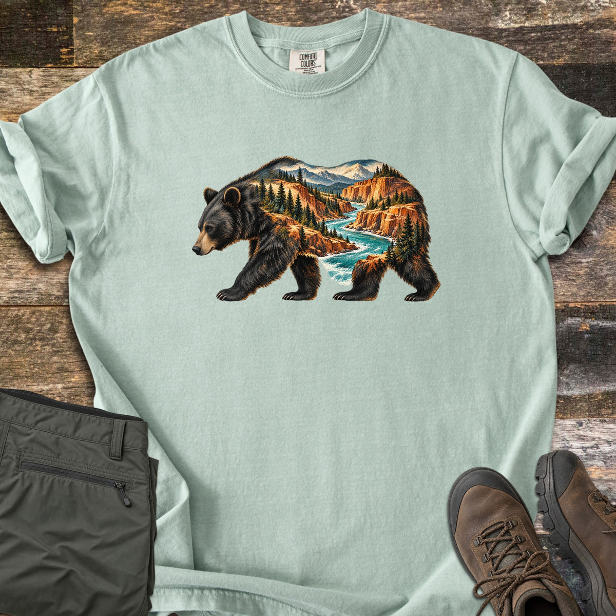 Bear River Canyon Comfort Colors T-shirt in Bay, laid flat. A walking black bear whose body is filled with a canyon scene of orange cliffs, pine trees, a turquoise river and snowy peaks. The sleeves are cuffed, with olive hiking pants and brown boots on weathered wood.