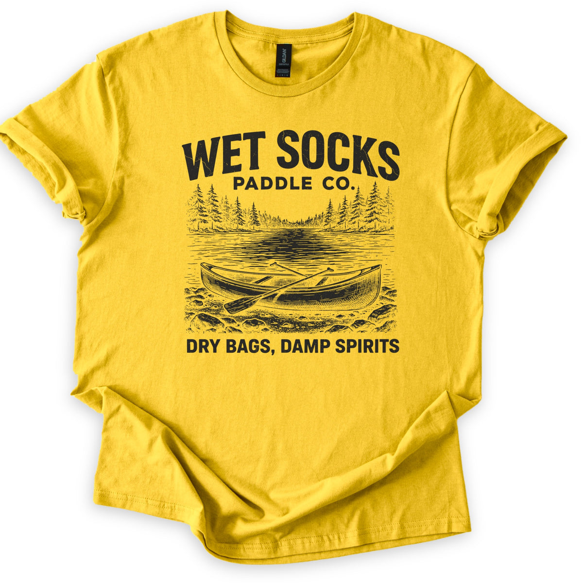 Wet Socks Paddle Co Comfort Colors T-shirt in Daisy, laid flat. A black etched drawing of a canoe with crossed paddles on a rocky shore, with a lake and pine trees behind, under the words Wet Socks Paddle Co. and above Dry Bags, Damp Spirits. White background.
