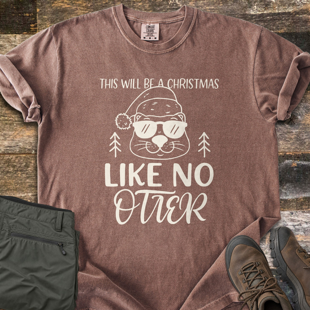 Christmas Like No Otter Comfort Colors T-shirt in Espresso, laid flat. A cartoon otter in a Santa hat and sunglasses sits between two pine trees, with This Will Be a Christmas above and Like No Otter below, in cream. Olive cargo pants and brown hiking boots lie on weathered wood.