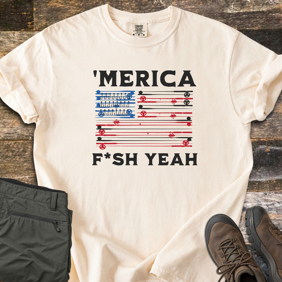Fish America Fish Yeah Comfort Colors T-shirt in Ivory, laid flat. Black text reads 'MERICA above an American flag made of red and black fishing rods, with rows of hooks in the blue corner, then F*SH YEAH. Folded olive pants and brown hiking boots sit on weathered wood.