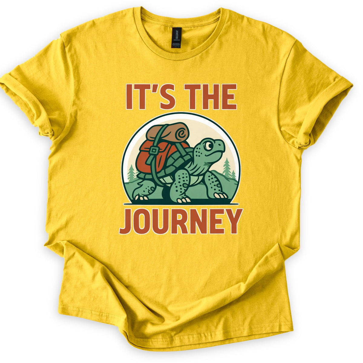 Slow Turtle Hiker Journey Comfort Colors T-shirt in Daisy, laid flat. A green cartoon turtle carrying a red backpack with a rolled sleeping bag walks past pine trees, framed by the words It's the Journey in rust-orange letters. Sleeves are cuffed on a white background.