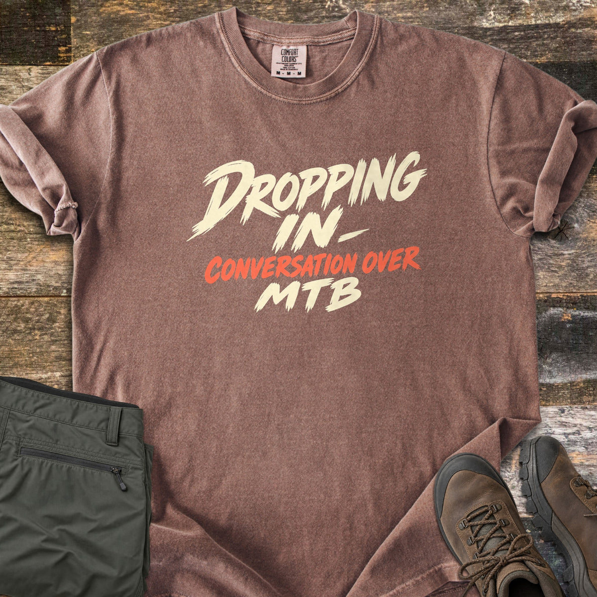 Dropping In Conversation Over Comfort Colors T-shirt in Espresso, laid flat. Cream brush-stroke letters read Dropping In- and MTB, with Conversation Over in orange-red between them. The shirt lies on weathered wood planks beside folded olive pants and brown hiking boots.