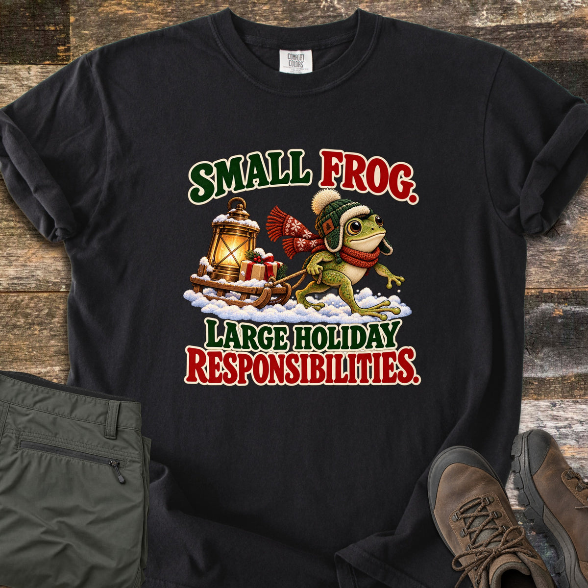 Small Frog. Large Holiday Responsibilities Comfort Colors T-shirt in Black, laid flat. A frog in a knit trapper hat and red scarf pulls a snowy wooden sled with a glowing lantern and gift, between the words Small Frog. Large Holiday Responsibilities. Olive pants and brown boots lie on wood planks.
