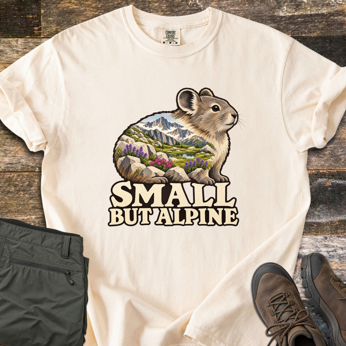 Small But Alpine Comfort Colors T-shirt in Ivory, laid flat. A pika whose body is filled with a mountain scene of snowy peaks, a lake, and purple and pink wildflowers among rocks sits above the words Small But Alpine. Green hiking pants and brown boots rest on weathered wood.
