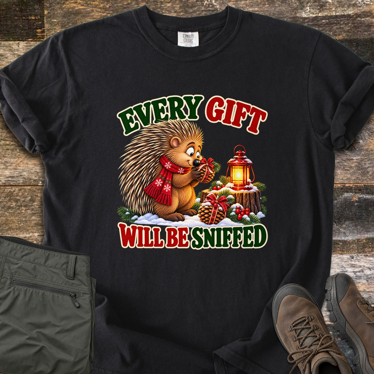 Porcupine Sniff Test Comfort Colors T-shirt in Black, laid flat. A cartoon porcupine in a red snowflake scarf sniffs a gift-wrapped pinecone beside a glowing lantern on a snowy stump, with the words Every Gift Will Be Sniffed. Green pants and brown hiking boots lie on weathered wood.