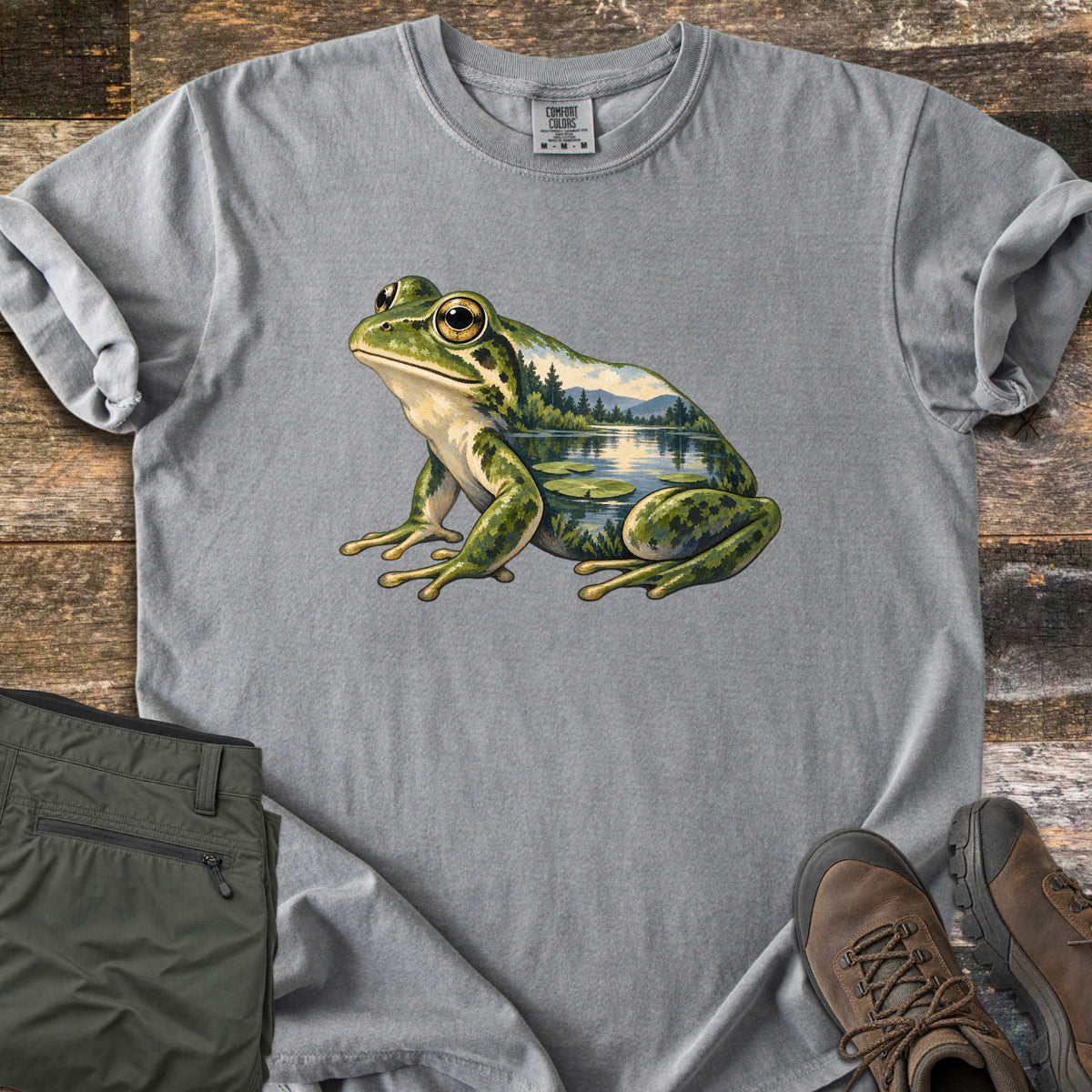 Lily Pond Frog Comfort Colors T-shirt in Grey, laid flat. A painted green frog sits in profile, its body filled with a lake scene of pine trees, mountains, and lily pads. Folded olive pants and brown hiking boots lie beside it on weathered wood boards.