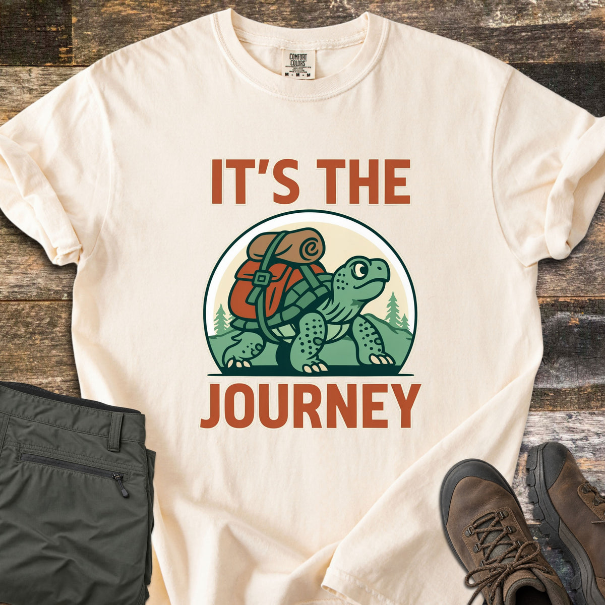 Slow Turtle Hiker Journey Comfort Colors T-shirt in Ivory, laid flat. A cartoon turtle carrying a red backpack and bedroll walks past pine trees and hills between the words It's the Journey. Olive hiking pants and brown hiking boots sit beside it on rustic wood planks.