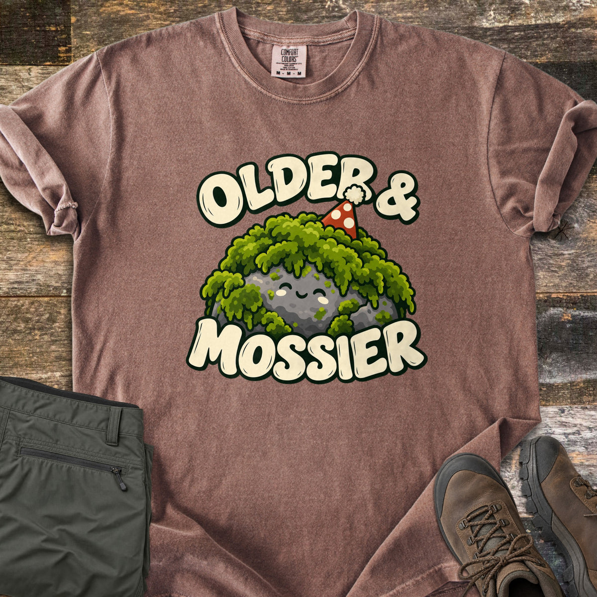 Older & Mossier Comfort Colors T-shirt in Espresso, laid flat. Cream letters read Older & Mossier around a smiling gray rock draped in green moss with a red polka-dot party hat. The shirt lies on weathered wood beside folded olive hiking pants and brown hiking boots.