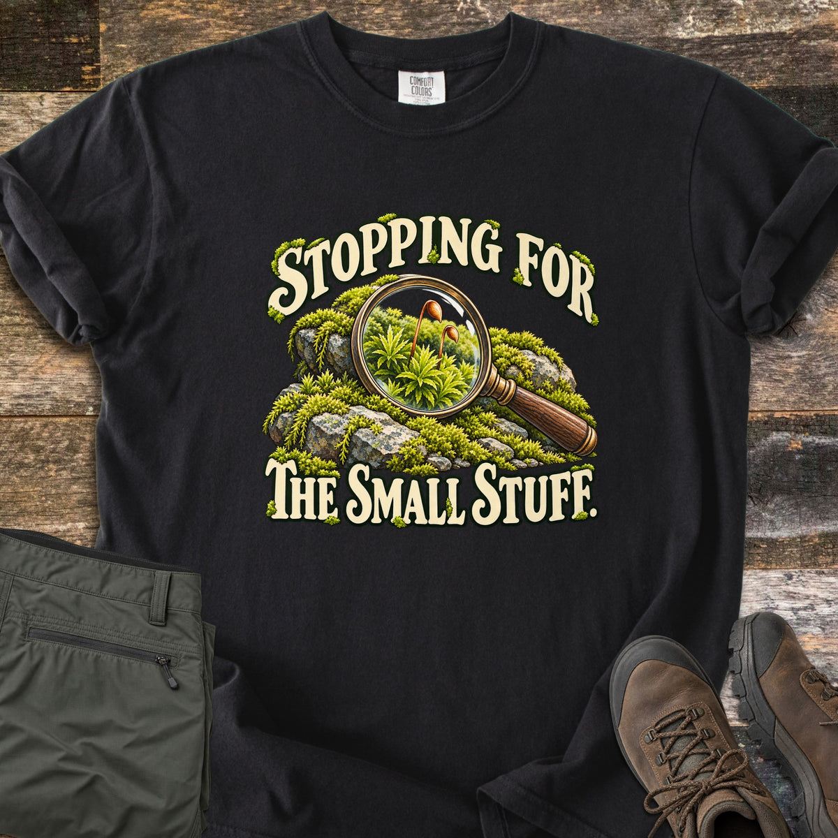 Stopping for the Small Stuff Comfort Colors T-shirt in Black, laid flat. A magnifying glass rests on mossy rocks, showing tiny moss sprouts, with the words Stopping for the Small Stuff. On rustic wood beside folded olive hiking pants and brown hiking boots.