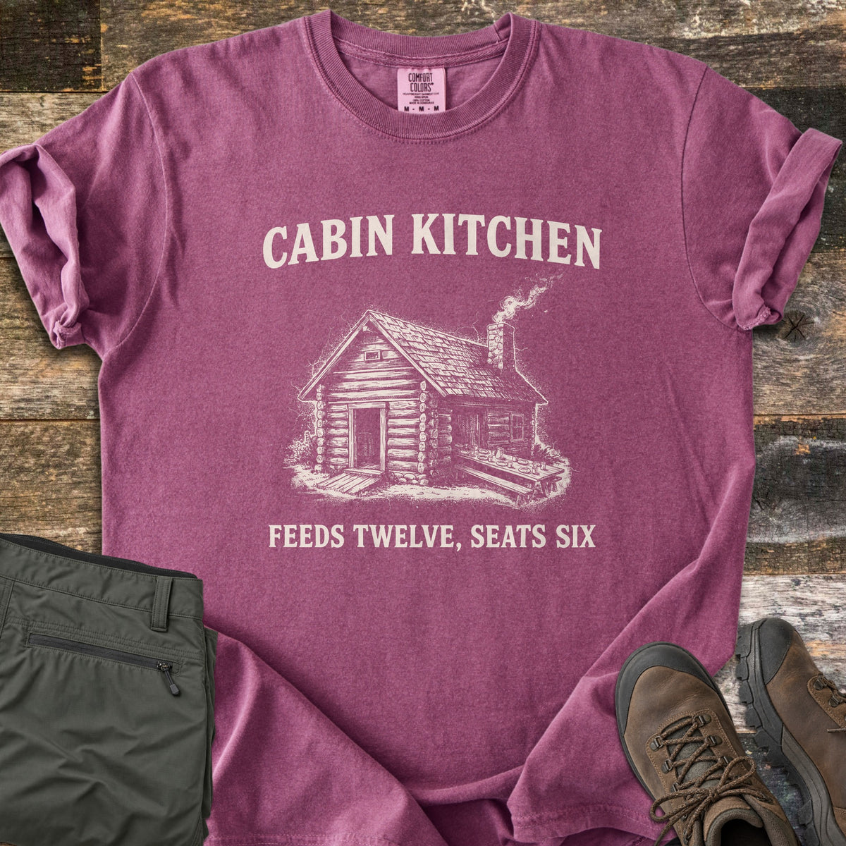 Cabin Kitchen Comfort Colors T-shirt in Berry, laid flat. An off-white sketch of a smoking log cabin with a long outdoor table sits between the words Cabin Kitchen and Feeds Twelve, Seats Six. Olive hiking pants and brown hiking boots lie beside it on weathered wood.