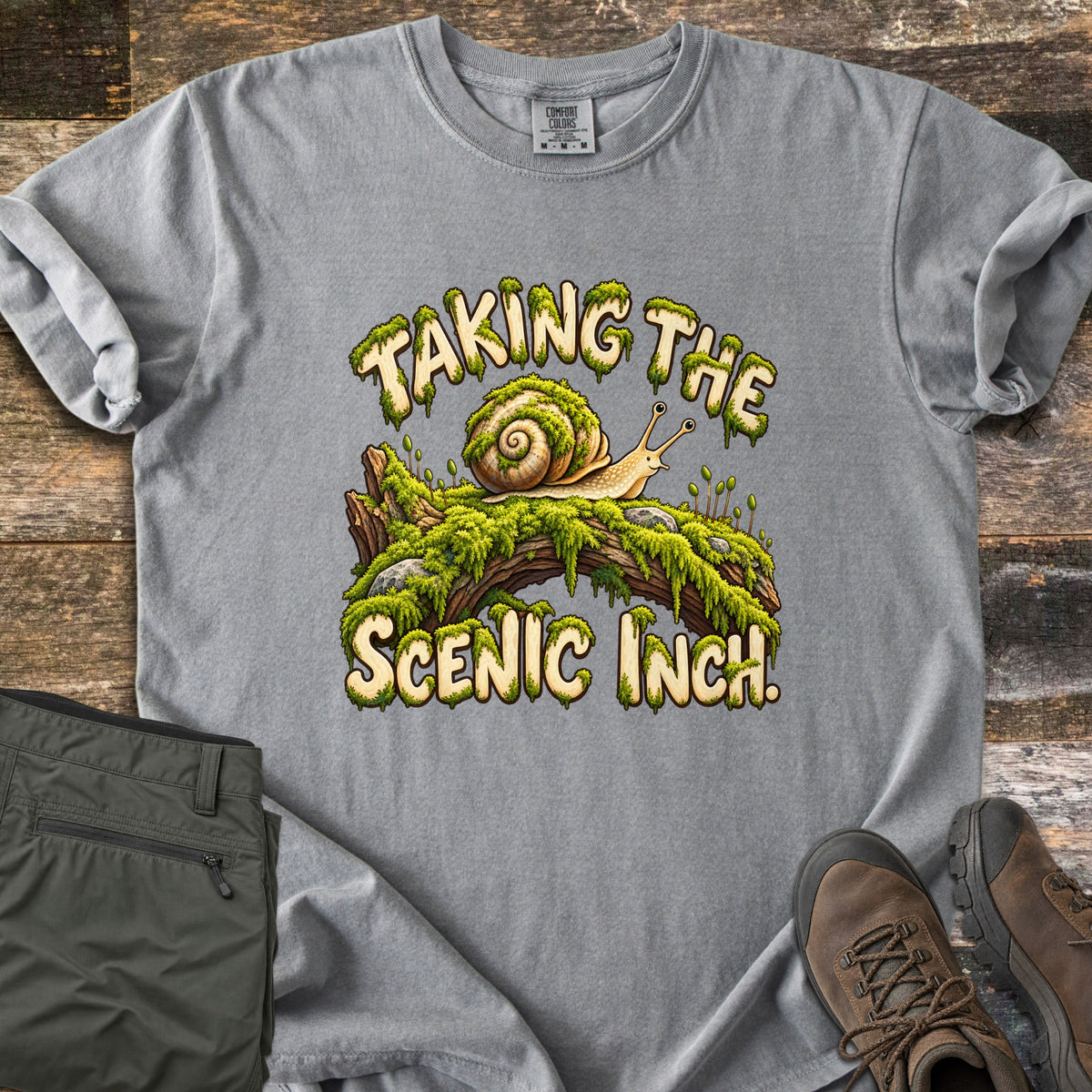 Taking the Scenic Inch Comfort Colors T-shirt in Grey, laid flat. A smiling cartoon snail with a spiral shell crawls along a mossy log between the words Taking the Scenic Inch., lettered in cream with dripping moss. Olive hiking pants and brown boots sit on a weathered wood floor.