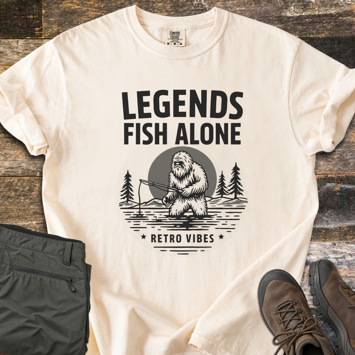 Legends Fish Alone Comfort Colors T-shirt in Ivory, laid flat. Black print of the words Legends Fish Alone above a Bigfoot fishing in a lake before a gray moon and pines, with Retro Vibes between two stars below. Olive pants and brown hiking boots lie on weathered wood.