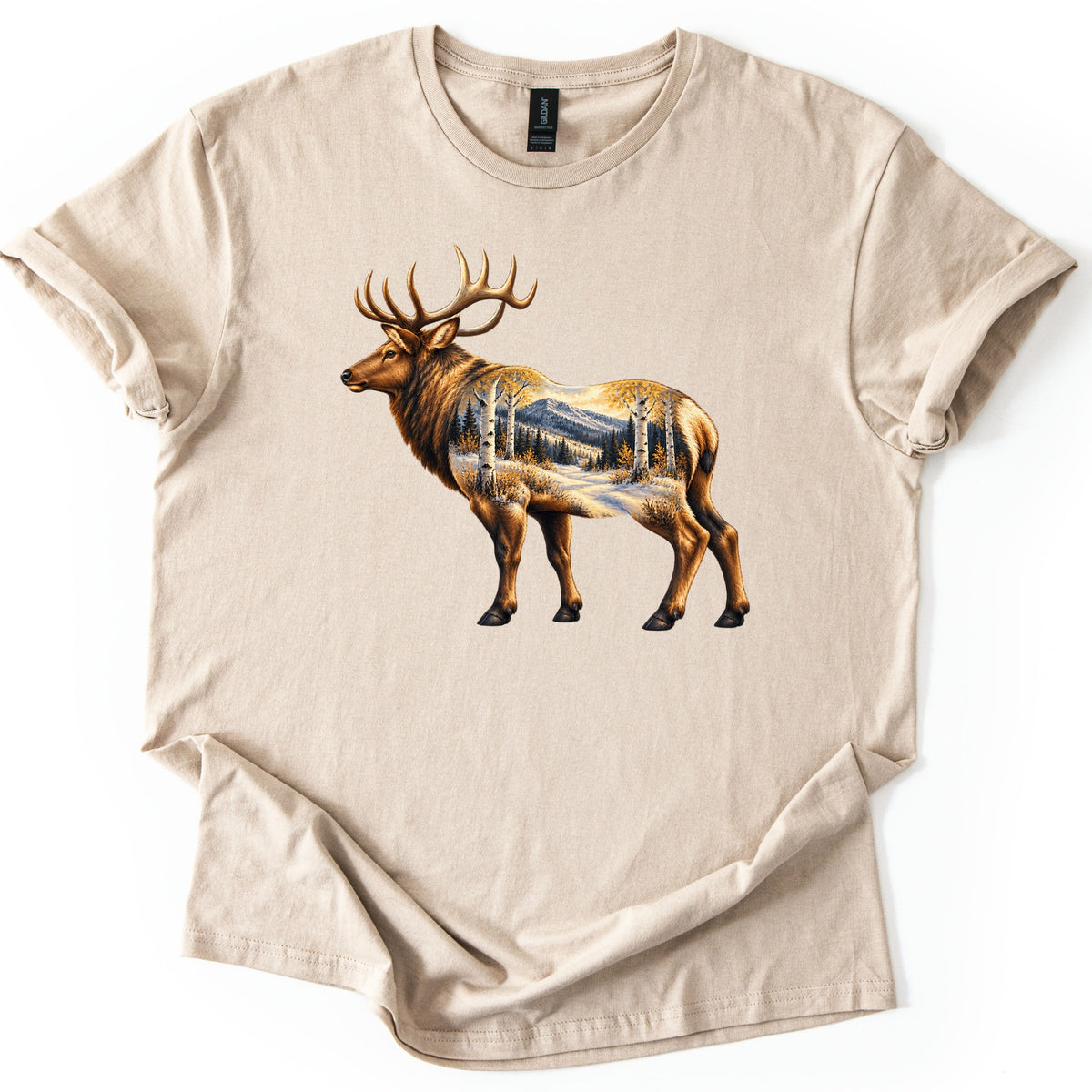 Aspen Pass Elk Comfort Colors T-shirt in Sand, laid flat. A realistic bull elk stands facing left, its body filled with a snowy mountain trail lined with white aspens in golden leaves and dark pines. No words. The shirt lies on white, hem folded up.