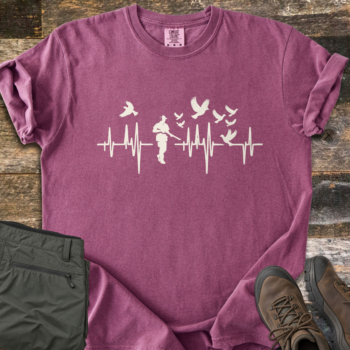 Bird Hunter Comfort Colors T-shirt in Berry, laid flat. A cream heartbeat line crosses the chest with a hunter silhouette in a cap holding a shotgun and birds flying up from the line. Folded olive pants and brown hiking boots lie beside it on weathered wood.