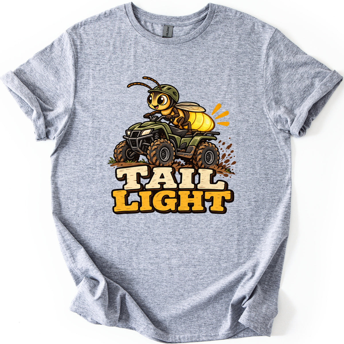 Tail Light Comfort Colors T-shirt in Sport Grey, laid flat. A cartoon firefly in a green helmet rides a muddy green ATV, its glowing yellow tail lit up, above the words Tail Light. The shirt lies on a white background with the sleeves rolled.