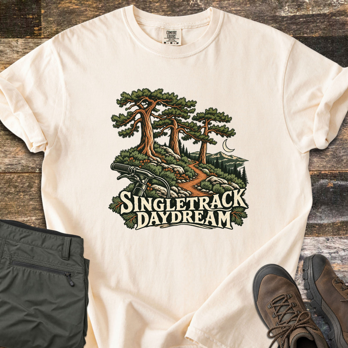 Singletrack Daydream Comfort Colors T-shirt in Ivory, laid flat. Three tall pines tower over a dirt trail winding past rocks toward mountains and a crescent moon, with bike handlebars in front and the words Singletrack Daydream. Set on wood with olive pants and brown hiking boots.