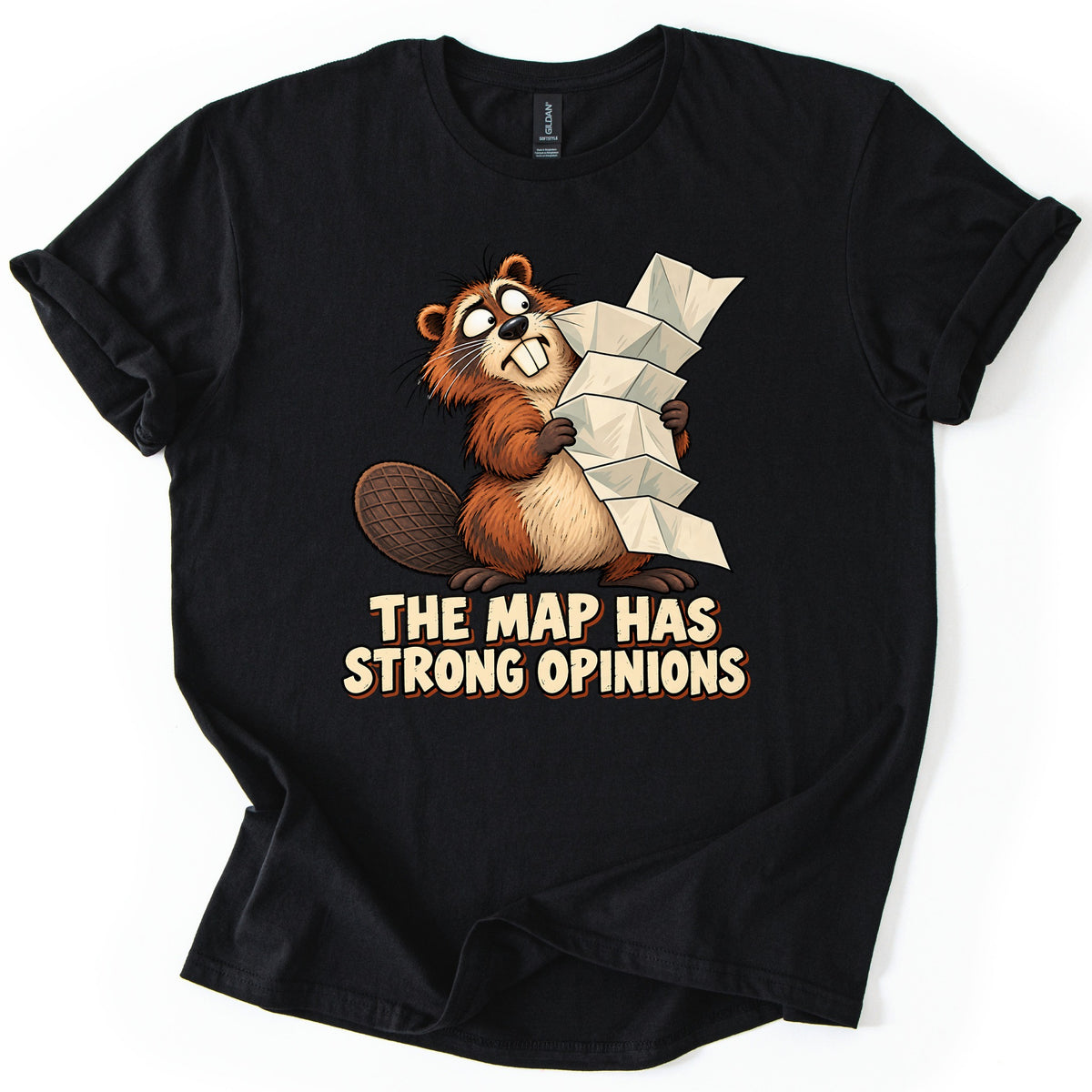 The Map Has Strong Opinions Comfort Colors T-shirt in Black, laid flat. A frazzled cartoon beaver with buck teeth hugs a towering stack of folded paper map above the words The Map Has Strong Opinions in cream lettering. The shirt lies on a white background with sleeves rolled.