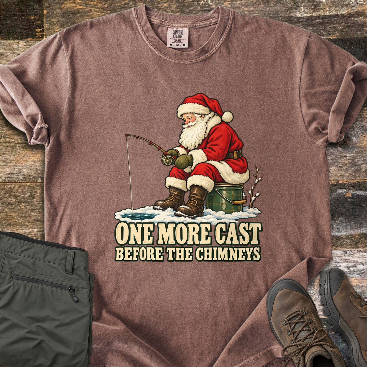 One More Cast Before the Chimneys Comfort Colors T-shirt in Espresso, laid flat. Santa in his red suit and brown boots sits on a green bucket, fishing through a hole in the ice, above the words One More Cast Before the Chimneys. Olive pants and hiking boots rest on weathered wood.