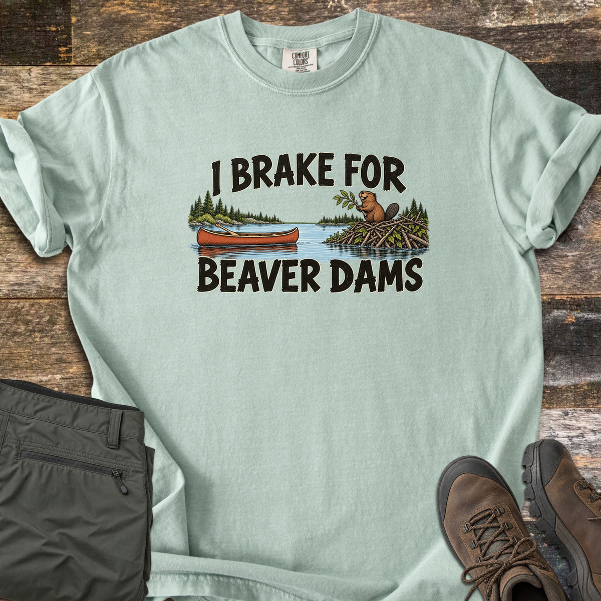 Brake For Beaver Dams Comfort Colors T-shirt in Bay, laid flat. The words I Brake For Beaver Dams frame a lake scene with a red canoe and a beaver on its stick dam among pine trees. Sleeves rolled, on weathered wood with folded green hiking pants and brown hiking boots.