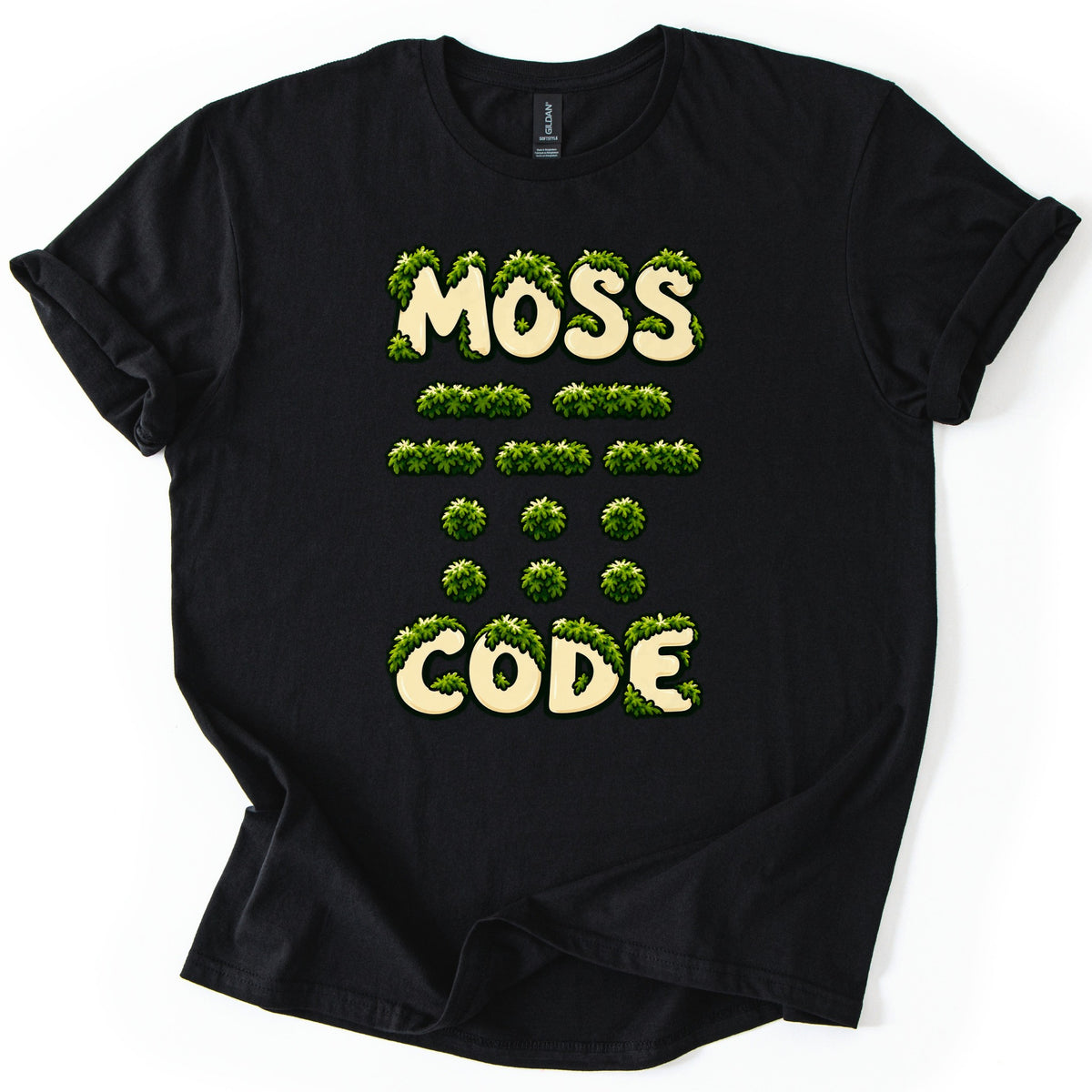 Moss Code Comfort Colors T-shirt in Black, laid flat. Cream bubble letters reading MOSS and CODE are overgrown with green moss, with rows of moss-covered Morse code dashes and dots between them. The shirt has rolled sleeves on a white background.