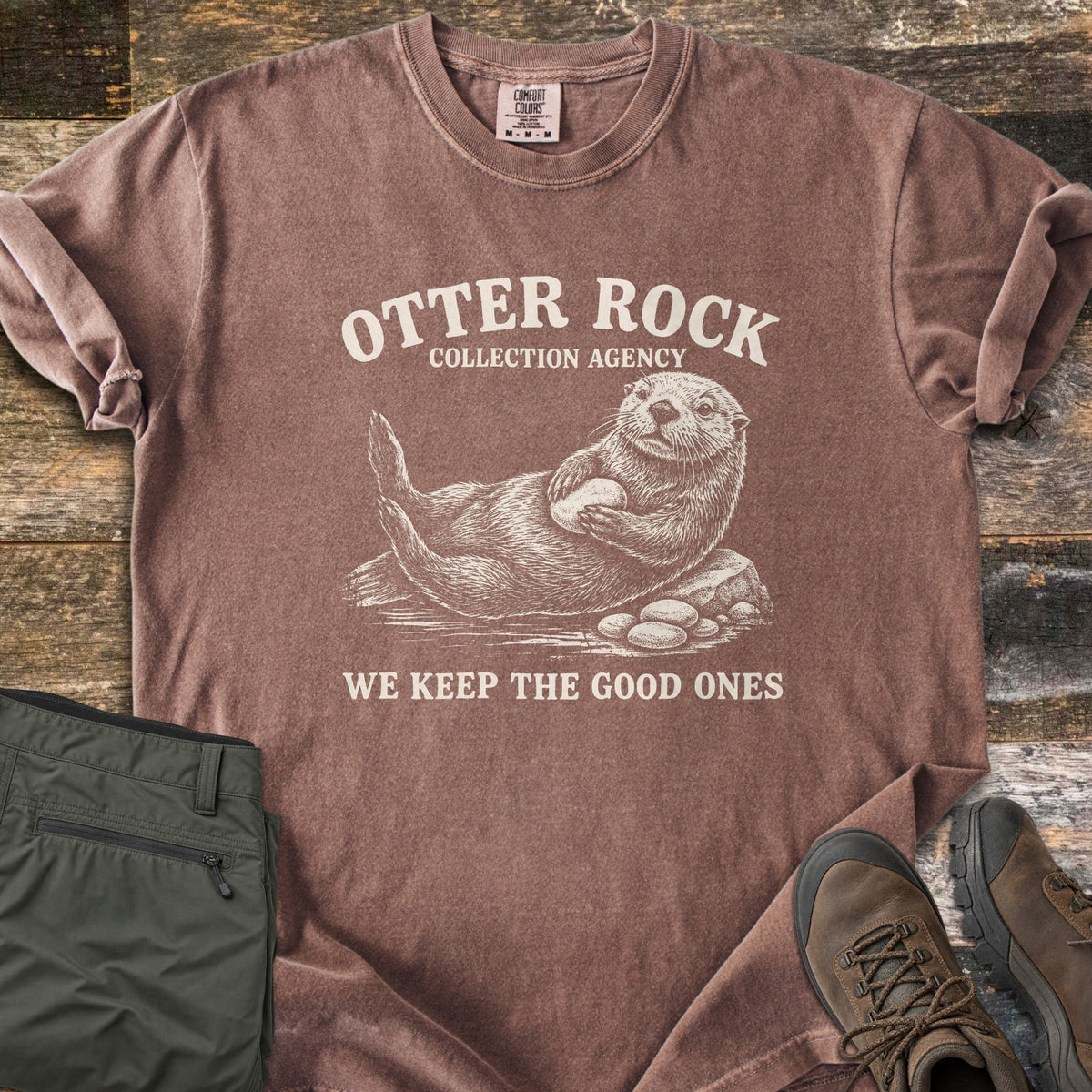 Otter Rock Collection Agency Comfort Colors T-shirt in Espresso, laid flat. A cream sketch of a sea otter lying on a rock holding a stone, under the words Otter Rock Collection Agency, with We Keep the Good Ones below. Shown on wood planks with olive pants and brown hiking boots.