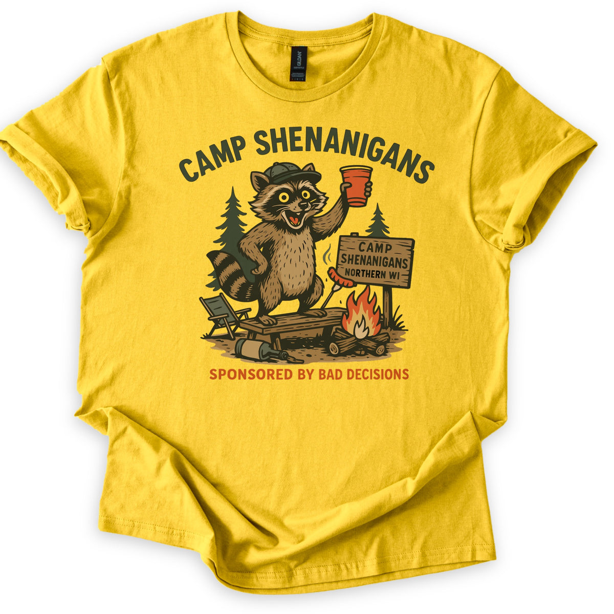 Camp Shenanigans Comfort Colors T-shirt in Daisy, laid flat. Camp Shenanigans arches over a raccoon in a cap toasting a red cup beside a campfire, pine trees and a sign reading Camp Shenanigans Northern WI, with Sponsored by Bad Decisions below. Sleeves cuffed, on white.