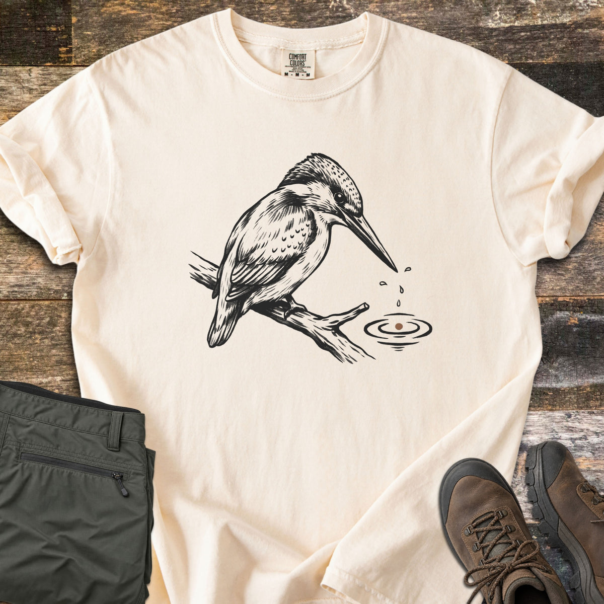Kingfisher Comfort Colors T-shirt in Ivory, laid flat. A black ink sketch of a kingfisher perched on a branch, looking down at water drops falling from its beak into rippling water, with no words. Olive pants and brown hiking boots sit beside it on weathered wood.