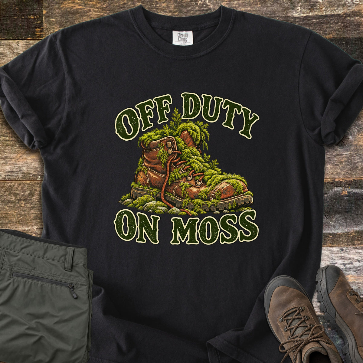 Off Duty on Moss Comfort Colors T-shirt in Black, laid flat. A worn brown hiking boot overgrown with green moss sits between the words Off Duty and On Moss in dark green letters. Laid on weathered wood with folded olive hiking pants and brown hiking boots.