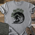 Surf Squatch Comfort Colors T-shirt in Grey, laid flat. A black silhouette of Bigfoot surfing inside a curling wave, with green palm trees behind, and no words. The shirt lies on weathered wood with folded olive pants and brown hiking boots.