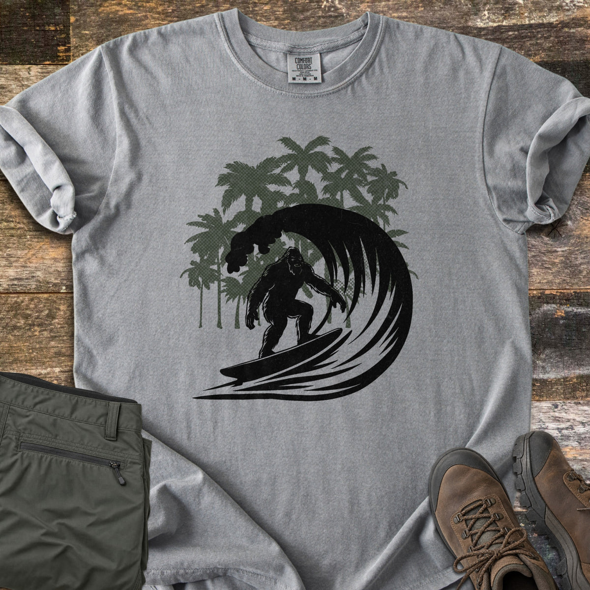 Surf Squatch Comfort Colors T-shirt in Grey, laid flat. A black silhouette of Bigfoot surfing inside a curling wave, with green palm trees behind, and no words. The shirt lies on weathered wood with folded olive pants and brown hiking boots.