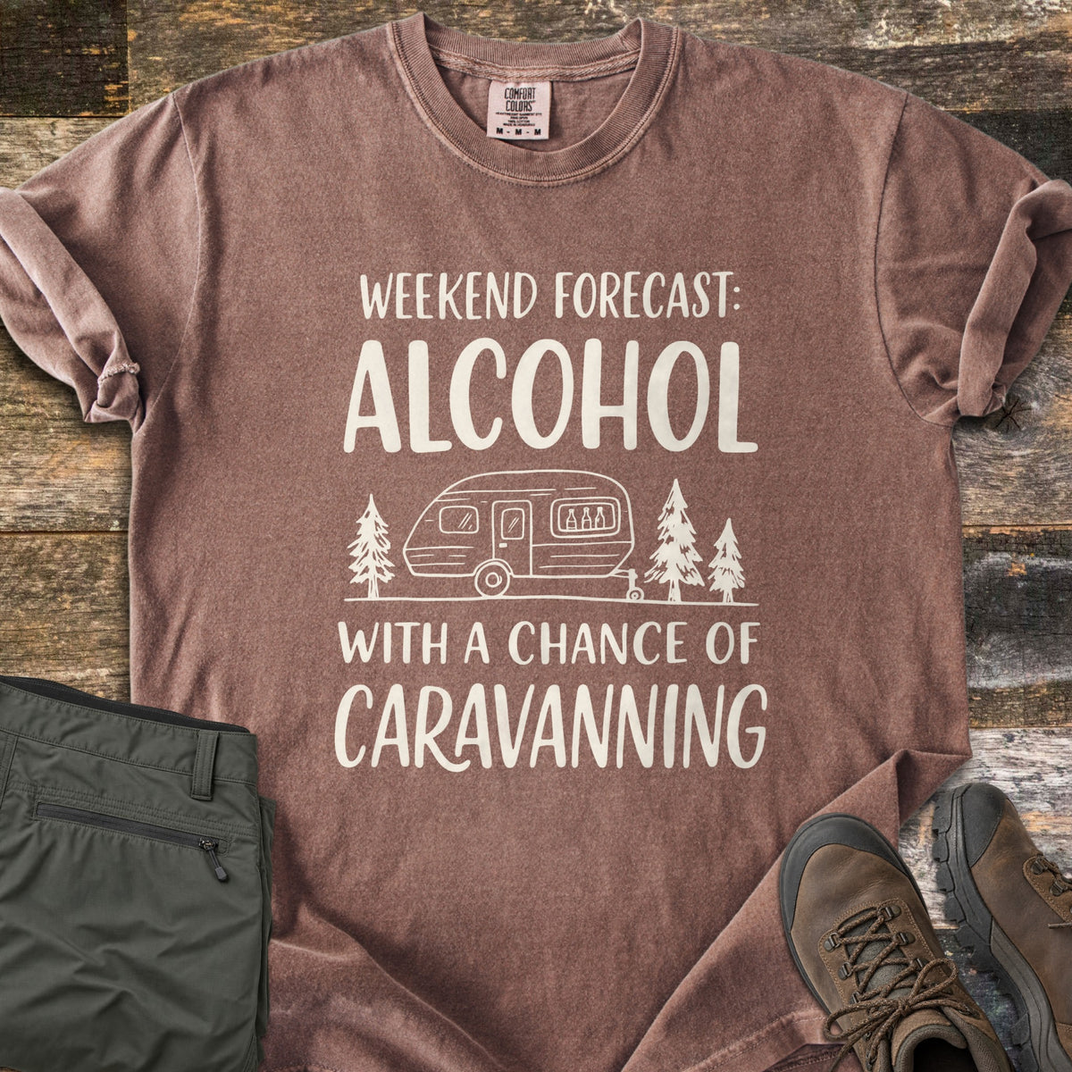 Alcohol With a Chance of Caravanning Comfort Colors T-shirt in Espresso, laid flat. Cream print reads Weekend Forecast: Alcohol above a caravan with bottles in its window between pine trees, then With a Chance of Caravanning. Olive trousers and brown hiking boots sit on dark wood planks.