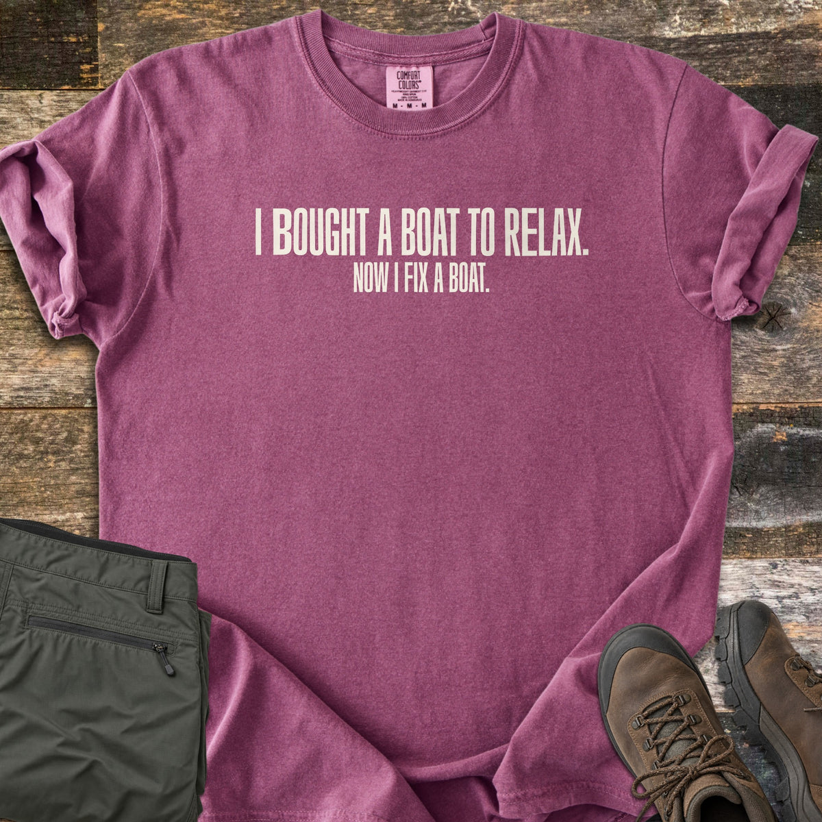 Boat To Relax Comfort Colors T-shirt in Berry, laid flat. Cream block letters read I BOUGHT A BOAT TO RELAX. with NOW I FIX A BOAT. in smaller type below. Sleeves are cuffed, with folded olive cargo pants and brown hiking boots on weathered wood planks.