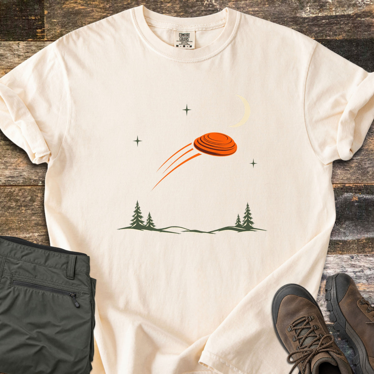 Moonrise Clay Comfort Colors T-shirt in Ivory, laid flat. An orange clay target flies past a pale crescent moon and small stars above green pine trees and hills, with no words. Folded olive cargo pants and brown hiking boots sit beside it on rustic wood planks.
