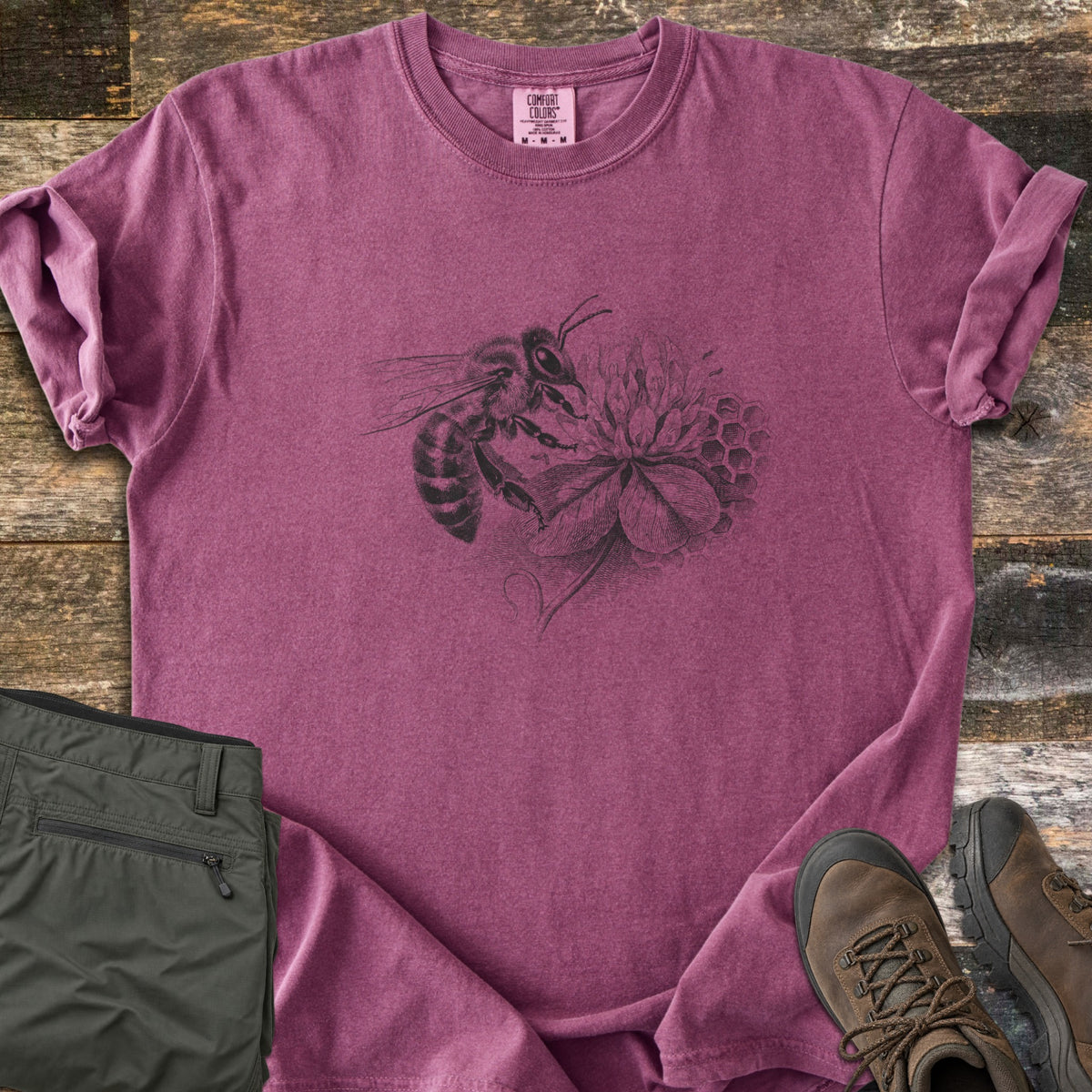 Honeybee On Clover Comfort Colors T-shirt in Berry, laid flat. A detailed black engraving-style illustration of a honeybee on a clover blossom with honeycomb cells behind it, no words. Sleeves rolled, on weathered wood planks beside olive hiking pants and brown hiking boots.