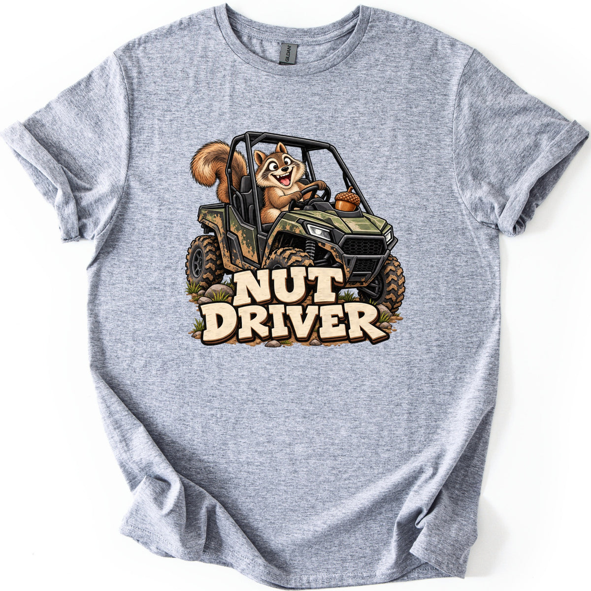 Nut Driver Comfort Colors T-shirt in Sport Grey, laid flat. A grinning cartoon squirrel drives a muddy camo side-by-side UTV over rocks and grass, an acorn on the hood, above the words Nut Driver in cream letters. The shirt lies on a plain white background.