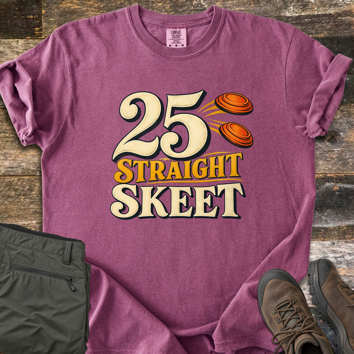 25 Straight Skeet Comfort Colors T-shirt in Berry, laid flat. Large cream numbers 25 with two orange clay targets flying beside them, above the words Straight Skeet in gold and cream. Sleeves are rolled on a wood plank surface with olive cargo pants and brown hiking boots.